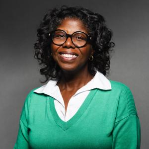Robin Sundaramoorthy wearing a green sweater and white collared shirt smiling for her headshot with a black backdrop.